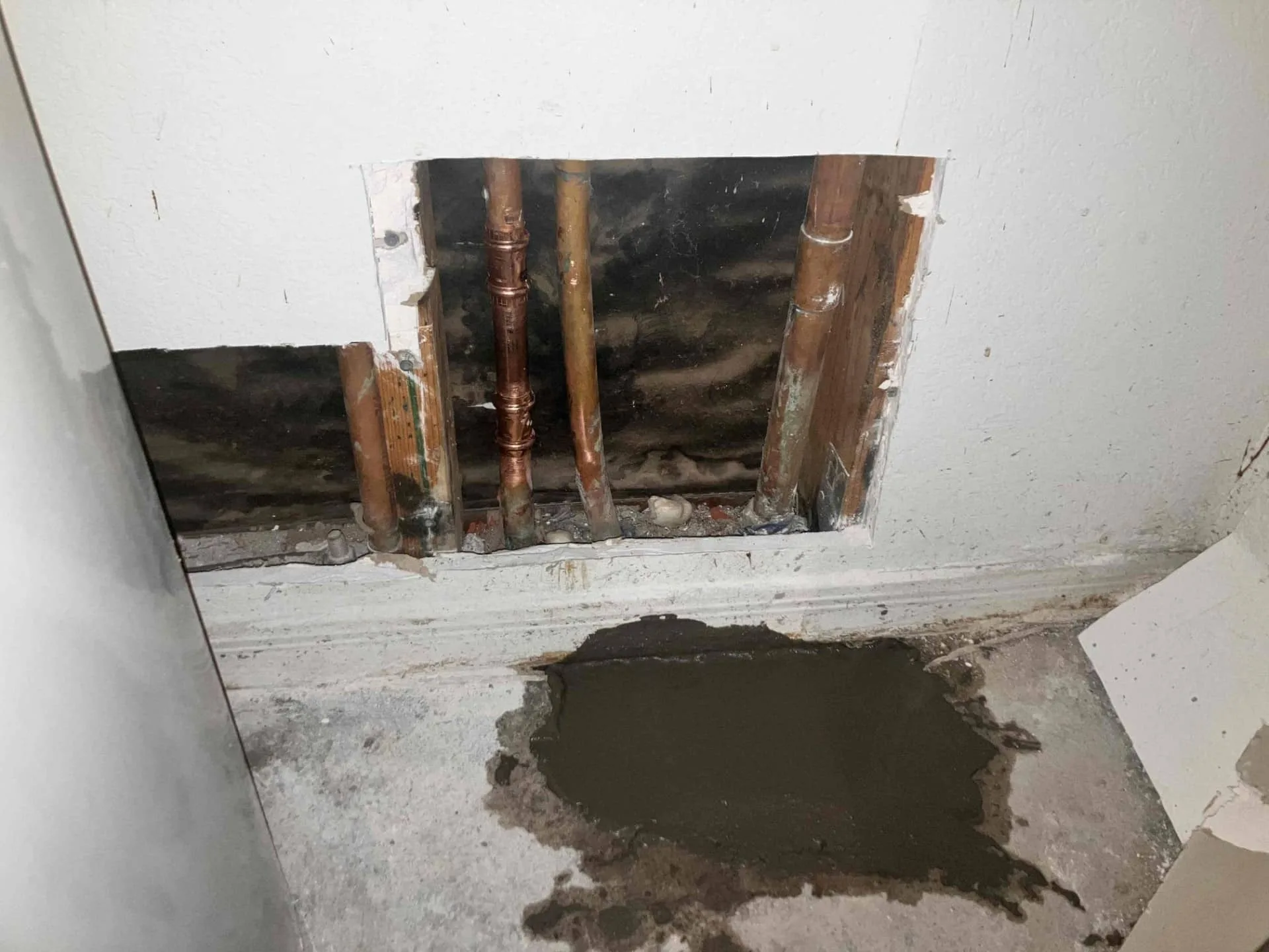 Exposed plumbing pipes behind a wall with fresh concrete patch on the floor, illustrating home repiping issues relevant to plumbing maintenance in Arizona.